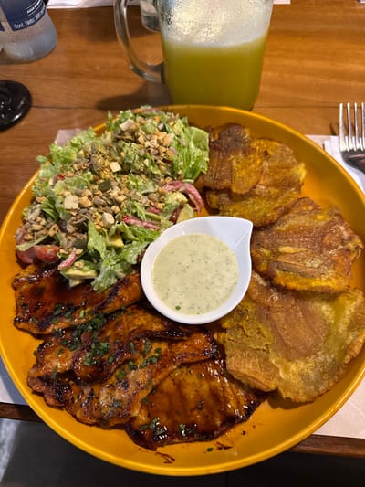 The scene depicts a meal served on a wooden table within a dining establishment located in the city of AMCO, Area Metropolitana Centro Occidente, Colombia. A large, yellow, oval-shaped plate occupies the majority of the frame, laden with food. On the left side of the plate are several pieces of grilled meat, possibly chicken or pork, coated in a dark, glossy sauce and topped with chopped green herbs. Adjacent to the meat is a substantial portion of green lettuce salad, mixed with diced red and green vegetables, and heavily garnished with a blend of seeds and chopped nuts. A small, white, ceramic boat-shaped dish, containing a light green, creamy sauce, is positioned between the meat, salad, and the third food component. The right side of the plate holds multiple golden-brown, flattened fried plantains (patacones).
In the upper left background, a clear plastic water bottle with "AGUA PURA" and "Cont. Neto: 300 ml" text is visible. To its right, a clear glass pitcher with a handle contains a light green, frothy beverage. A silver fork is partially visible on the far right edge of the frame, resting next to a white napkin or menu on the wooden table. No individuals are present in the image.