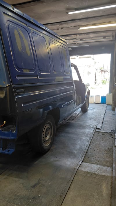 The image depicts the interior of a garage in Belovo, Russia. A dark blue van, with its driver's side door open and the rear partially disassembled, is the central subject. The van appears to be undergoing maintenance or repair, evidenced by the exposed wiring at the rear and patches of primer on the side panels. The garage floor is made of concrete, with some areas covered by wooden planks. Fluorescent lighting fixtures are mounted on the ceiling, illuminating the space. Outside the garage, through an open doorway, several plastic containers and a brick building are visible. The overall scene suggests a functional, working garage environment.