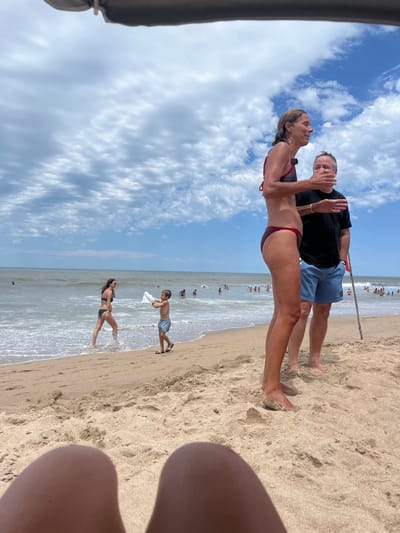 The image captures a lively beach scene on a sunny day. In the foreground, slightly to the left, a couple stands on the sandy shore. The woman, wearing a dark red or maroon bikini, has her head tilted up, looking towards the man. She has long hair and appears to be smiling or talking. The man stands beside her, dressed in a black t-shirt and light blue shorts. His right hand is raised, possibly in a gesture of talking or waving, and he is looking towards the woman.
The beach itself is expansive, with light-colored sand. The ocean is visible behind the couple, with gentle waves breaking near the shoreline. Numerous people are scattered in the mid-ground and background, both in the water and on the sand, indicating a busy beach environment. Closer to the water's edge, in the mid-ground, a young person (possibly a boy) in light-colored shorts is seen standing, holding a white object. Further out, another woman in a dark swimsuit is standing in the shallow water.
The sky above is bright blue with scattered white, fluffy clouds. The overall weather appears clear and pleasant, suggesting ideal conditions for a beach outing. On the far left of the frame, a dark, blurry object, possibly part of a beach umbrella or tent, is partially visible, indicating the photographer may be under shade. On the far right, a blurry human leg or thigh is partially visible, suggesting the photo was taken from a low angle, possibly by someone sitting or lying on the sand. No discernible text is visible in the image.