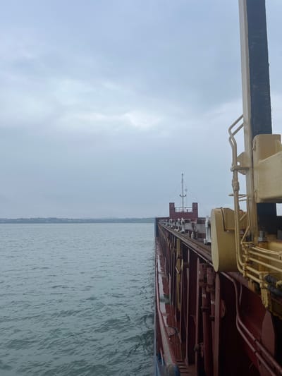 The image depicts the starboard side of a large maritime vessel, likely a cargo ship, situated in coastal waters under an overcast sky. The location is near Bangor, United Kingdom, with a distant landmass visible across the horizon.
In the immediate foreground, to the right, a tall, beige-colored structural element, possibly part of a crane or the ship's superstructure, features a prominent vertical black strip and yellow hydraulic piping with associated mechanical components. The vessel's hull, stretching from the mid-ground towards the distant horizon on the right, is predominantly dark red or maroon, with some lighter blue sections visible near the waterline. The hull displays numerous vertical ribs or supports, and reflections of the grey sky are present on its wet surface. Along the ship's deck, a series of regularly spaced white and grey fittings, possibly stanchions, lashing points, or cargo gear components, are visible. Further along the vessel's deck, a mast or antenna structure with radar domes is discernible against the distant horizon.
The water surface to the left of the vessel exhibits minor ripples, indicating calm to light choppy conditions. The sky is uniformly grey and heavily clouded, suggesting an overcast day. A low-lying landmass, appearing as a dark green-grey outline of trees and possibly some structures, stretches across the background horizon. No individuals are visible in the scene.