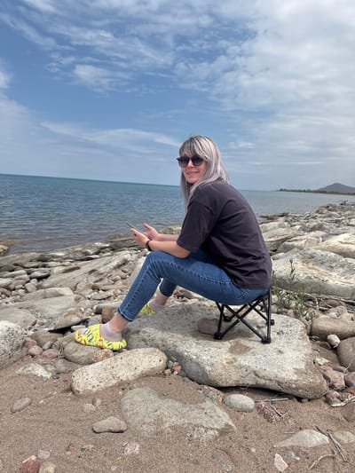 A woman with blonde hair and sunglasses sits on a portable black chair on a rocky shore beside a body of water. She is wearing a dark t-shirt, blue jeans, and yellow shoes with a cartoon pattern. The woman is holding a smartphone in her hands. The water is calm and appears to be a lake. The shore is covered in rocks and sand. In the background, there is a distant shoreline with hills and mountains under a partly cloudy sky. The scene is likely located near the city of Tong, Kyrgyzstan, which is situated on the southern shore of Issyk-Kul Lake.