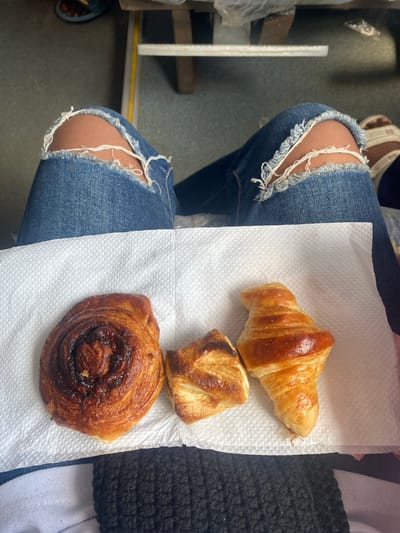 The image shows a person sitting down, wearing ripped jeans and shoes. On a white napkin placed on their lap are three pastries: a croissant, a square-shaped pastry, and a spiral pastry. The scene is likely inside a vehicle, based on the flooring and a glimpse of a window. There is no direct evidence to determine if the scene is in Cotonou, Benin.