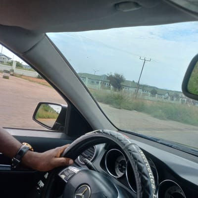 The image, taken from inside a Mercedes-Benz vehicle, shows a person's left hand on the steering wheel. The hand is wearing a gold watch with a black leather band. The steering wheel is covered with a black, diamond-patterned cover. The dashboard displays gauges and air vents. Outside the car, a road is visible, with houses and a fence lining the side. In the distance, power lines and poles are present, along with a cloudy sky. The side mirror reflects a grassy embankment. The location is identified as Dorowa, Nigeria.