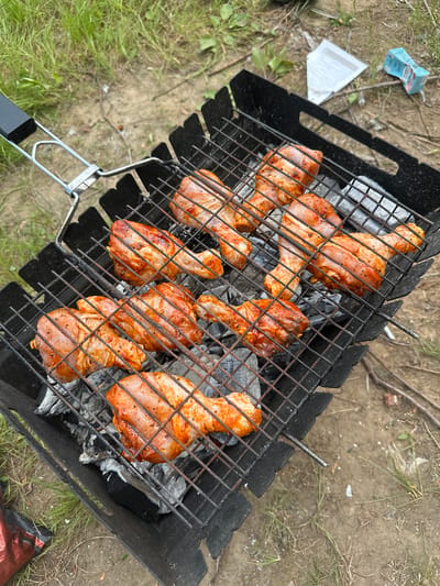 In an outdoor setting with dry, sandy soil and sparse grass, a black metal grill is placed on the ground. The grill contains hot coals, and a wire rack is positioned above them. On the rack, several raw chicken drumsticks are arranged. The drumsticks are coated in a reddish-orange marinade and seasoned with black pepper. A silver metal handle is attached to the grill. In the background, there are some scattered white papers and a small blue and white box. The overall scene depicts food preparation for a barbecue. The location is identified as Pravda, Russia.