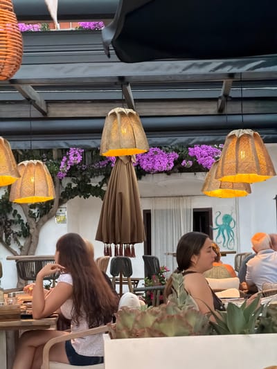 The image depicts an outdoor seating area of a restaurant, likely in Begur, Spain, given the presence of bougainvillea flowers. The scene is lit by several large, rustic lamps with woven shades, casting a warm glow. In the foreground, two women are seated at tables. The woman on the left, with long brown hair, is wearing a floral print top and denim shorts, and is looking towards the left. The woman on the right, also with brown hair pulled back, is wearing a dark top and appears to be looking forward. Behind them, other patrons are visible, including a man with a bald head wearing a white shirt and another person in the background. The restaurant setting includes wooden furniture, potted plants, and an octopus mural painted on a white wall. A closed, tan umbrella with tassels hangs in the center of the frame. The overall atmosphere suggests a relaxed evening meal.