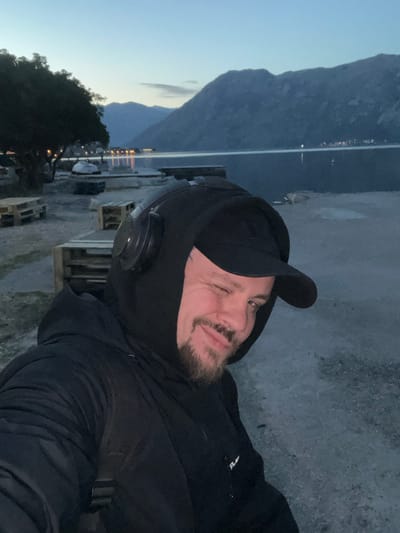 An outdoor, eye-level, slightly off-center portrait captures a male subject at dusk in Bogdašići, Montenegro. The male, positioned in the foreground, is winking with his right eye (viewer's left) and exhibiting a slight smile. He has a short beard and mustache, and a silver-colored hoop nose ring is visible on his left nostril. He is dressed in multiple layers of black clothing, including a baseball cap, a hooded sweatshirt, and a padded jacket. Black over-ear headphones are worn over the hood. A dark strap, possibly from a backpack, is visible over his left shoulder.
The background features a calm body of water, consistent with a bay or fjord, reflecting the ambient twilight sky. Distant mountains rise from the water's edge, silhouetted against the horizon. Lights from a distant settlement are visible on the far side of the water, creating reflections on its surface. The immediate shoreline consists of gravel or coarse dirt. To the left, dark trees with visible foliage are present. Several stacks of wooden pallets are discernible on the shore behind the subject, along with indistinct objects resembling small boats or covered structures near the water's edge. The overall lighting is dim, indicating late evening or early night.