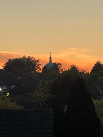 The image presents an evening scene at twilight or sunset, likely captured in High Wycombe, United Kingdom. The sky dominates the upper and left portions, displaying a warm gradient of colors. Starting from a pale yellow-orange at the top left, it transitions to a more vibrant and intense orange-red streak of clouds across the middle, suggesting the last light of the setting sun.
In the mid-ground, a distinctive architectural structure is visible, resembling a church or chapel. It features a light blue-green dome topped with a slender spire and a cross, silhouetted against the colorful sky. Surrounding this building and filling much of the foreground and right side are numerous trees with dense foliage, appearing as dark silhouettes due to the backlighting. The edges and uppermost branches of some trees are kissed by the orange light, creating a sparkling effect.
Below the treeline, dark silhouettes of residential buildings or houses can be discerned. On the far left, a window in one of these buildings emits a subtle, warm yellow light, suggesting an interior illumination. Faint power lines or cables are also visible traversing the upper right section of the sky. The overall scene conveys a serene and peaceful atmosphere typical of a quiet evening in an urban or suburban setting. No people or visible text are present in the image.
