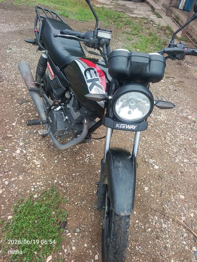 A black Keeway motorcycle is parked on a dirt and gravel ground. The motorcycle is facing forward and slightly to the right. It has a black seat, a black front fender, and a circular headlight with the "KEEWAY" logo below it. The motorcycle also has a black luggage rack on the back. In the background, there is some green grass and a concrete structure. The timestamp "2026/06/19 06:54" and the text "natha" are overlaid on the bottom left of the image. The scene appears to be outdoors, possibly in a rural or semi-urban area. No people are visible in the image.