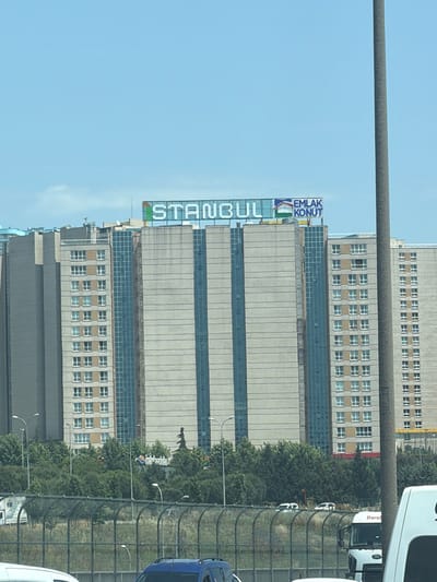 The image, taken in Avcılar, Turkey, captures a towering multi-story building under a clear blue daytime sky. The perspective is from a slightly elevated position, looking across a road or a raised highway towards the structure.
The central subject is a tall, modern high-rise building featuring a mix of light-colored concrete panels and dark, reflective glass sections. The top of the building is adorned with a prominent blue sign that reads "İSTANBUL" horizontally and "EMLAK KONUT" vertically on its right side. Below this main building, or possibly part of the same complex, are sections with numerous small, regularly spaced windows.
A thick, horizontal gray pole, likely part of a street light or overhead infrastructure, traverses the upper third of the frame. Below this pole, a line of dense green trees runs horizontally, separating the buildings from a road area. A tall, dark metal mesh fence is visible along the edge of the road, behind which several vehicles are partially visible. These include a white truck with "Dural" written on its side, a blue car, and parts of other white vehicles. The overall scene depicts a typical urban environment.