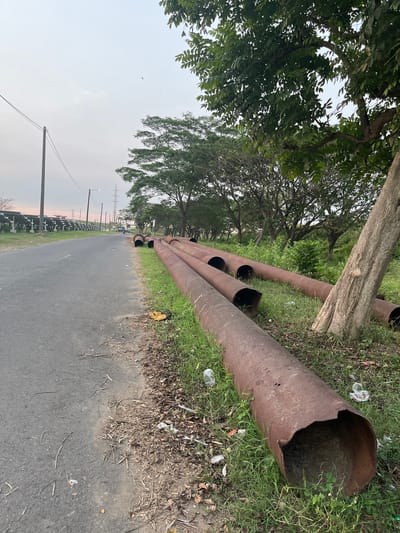 A street scene in Besuki, Indonesia, features a paved road with a grassy verge lined with several large, rusty pipes. The pipes are laid horizontally on the grass, appearing to be discarded or awaiting installation. A large tree trunk is visible next to the pipes, its bark textured and aged. Beyond the pipes, a line of smaller trees forms a dense, leafy backdrop. In the distance, power lines and transmission towers are visible, suggesting proximity to industrial or utility infrastructure. The sky is overcast, with a pale, diffused light indicating it is either early morning or late afternoon. There are no people prominently visible in the scene, though a motorcycle can be seen on the road in the distance, with what appears to be a small solar panel farm on the left side of the road. The overall impression is one of an undeveloped or transitional area within the city.