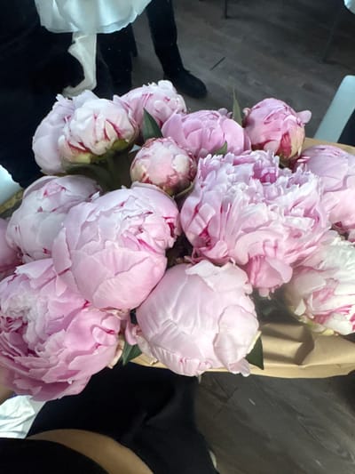 A close-up shot displays a bouquet of pink peonies, their petals varying in shade from pale pink to white with crimson streaks. The flowers are arranged in a brown paper wrapping. In the background, dark wooden floorboards are visible. To the left, a person wearing black clothing is partially seen, with white fabric, possibly part of a dress or shirt, draped over their shoulders. The scene is dimly lit, with some areas in shadow. The image does not provide any specific clues about the location in the city of Istra, Russia.