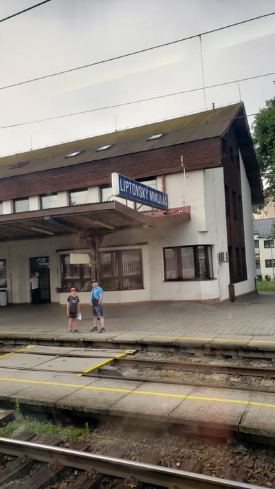 The image shows a train station platform in Liptovský Mikuláš, Slovakia. A sign on the building reads "LIPTOVSKÝ MIKULÁŠ". Two people, an adult male and a young boy, are standing on the platform facing the camera. The adult is wearing a blue t-shirt, denim shorts, and a black crossbody bag. The boy is wearing a dark t-shirt with an "Emirates" logo, dark shorts, and a red baseball cap. They are standing near the entrance of the station building, which has a covered walkway. Train tracks are visible in the foreground, separated from the platform by a concrete barrier with yellow lines. The sky is overcast.