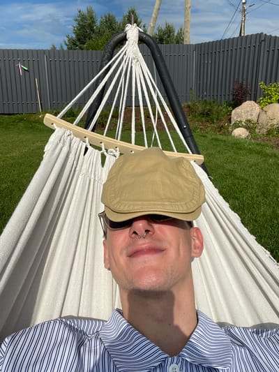 A young man with a septum piercing and a light brown flat cap is lying in a white hammock outdoors. He is wearing sunglasses that partially obscure his eyes and a blue and white striped button-up shirt. The hammock is suspended from a black metal stand, and the scene appears to be a backyard in Simagino, Russia. A grey corrugated metal fence is visible in the background, along with green grass, trees, and a bright blue sky with scattered clouds. The sun is shining, casting shadows.