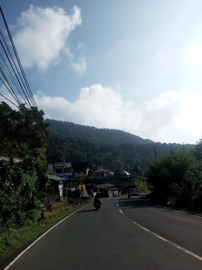 The image depicts a paved road leading uphill towards a densely forested mountain. A motorcyclist is traveling away from the viewer on the road. On the left side of the road, there's a building with the word "POLISI" visible, suggesting a police post. The surrounding area includes lush greenery and trees. Overhead, numerous power lines intersect the view. The sky is partly cloudy, with bright sunlight in the upper right corner. There are no identifiable details linking the image to the specific city or country mentioned.