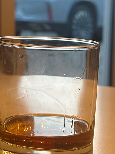 This is a close-up, eye-level, horizontal shot of a glass of amber liquid, likely beer or a similar beverage, on a table. The glass is partially filled, with condensation visible on the exterior, creating streaks and droplets. The liquid itself is translucent and reflects light, highlighting its color and some subtle bubbles within.
In the background, the scene is out of focus. To the left, a blurry silhouette of an object with a radial pattern is visible, resembling the front of a hairdryer or a fan. The rest of the background consists of a neutral, textured surface, possibly a wall or furniture, with areas of light and shadow. The overall lighting suggests an indoor setting, possibly during daylight hours given the brightness, but without strong directional light.
The focus is entirely on the glass and its contents, drawing attention to the texture of the condensation and the richness of the liquid. The image evokes a sense of casual enjoyment or relaxation, typical of consuming a drink. There is no visible text in the image. The scene is set in a domestic or casual environment, with no specific clues about the exact location within Zubovo, Russia, other than the provided context.