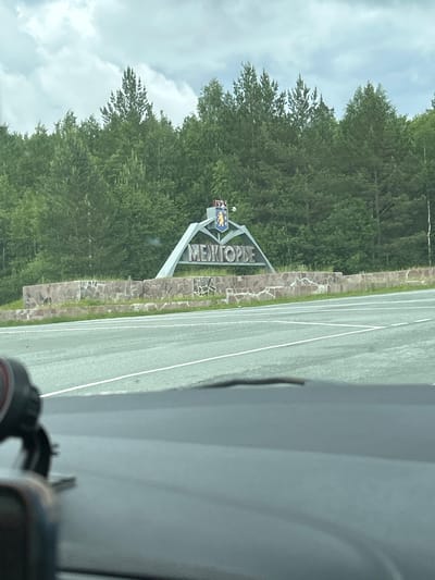 A frontal medium shot captures the entrance sign to the closed administrative-territorial formation "Mezhgorye", Russia, set against a backdrop of dense green forest and a cloudy sky. The sign, a large, angled structure in grey metal, displays the town's name in Cyrillic letters. Above the name, a coat of arms featuring a bear and a shield is visible. The sign is supported by a low, rough stone wall that extends horizontally across the frame. In the foreground, the grey asphalt of a road with white lane markings is prominent, suggesting the image was taken from a vehicle. The lower part of the frame is blurred, showing the interior of a car's dashboard, including a black object, possibly a camera or a dashcam. The scene is outdoors during the daytime, with overcast conditions.