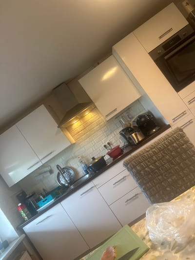 This is an indoor, medium-angle, eye-level shot of a modern, white kitchen. The kitchen features white cabinets with silver handles, a dark countertop, and a tiled backsplash. Appliances visible include a built-in oven with a digital display showing "18:57," a range hood with under-cabinet lighting illuminating the backsplash, a sink with a faucet, and various kitchenware on the countertop such as pots, a red bowl, a coffee maker, and what appears to be a blender or food processor. To the right of the frame, a textured grey fabric item, possibly a chair or cushion, is partially visible, along with a clear plastic bag containing items. In the foreground, a green cutting board with a knife and a piece of raw meat is on a floral-patterned surface. The lighting suggests indoor illumination, possibly from the under-cabinet lights and overhead ceiling light. The image's perspective is slightly tilted. The location is identified as a kitchen in Mainburg, Germany.