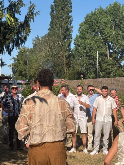 The image depicts a group of men gathered outdoors, with a clear blue sky and lush green trees in the background. The setting appears to be a backyard or park, given the presence of grass, a wooden fence, and rose bushes. The overall ambiance suggests a casual social gathering, possibly a barbecue or celebration.
In the foreground, a man is facing away from the camera, wearing a light-colored, textured, open-weave shirt and brown trousers. His presence dominates the immediate view, with his back turned to the viewer, obscuring some of the other individuals.
To his left, several men are visible. One man is wearing a blue and white patterned short-sleeved shirt, a straw hat, and sunglasses. Another man is in a white shirt and dark trousers. Further left, another man in a white shirt is partially visible.
To the right of the man in the foreground, a group of men are standing. One man in a white shirt and shorts is gesturing with his finger. Another man, wearing a light blue polo shirt, is shielding his eyes from the sun. Next to him, a man in a white short-sleeved shirt and light gray pinstriped trousers is smiling and holding a drink. To his right, a man with a colorful floral shirt and a lei around his neck is also visible.
The image was taken in Hem, France, and the specific location within Hem appears to be a private garden or outdoor event space. The lighting suggests it is daytime, likely late afternoon due to the warm, soft light and the long shadows cast on the grass. The men are dressed in a variety of casual attire, ranging from floral shirts to plain white shirts, with some wearing hats and sunglasses. The mood seems relaxed and convivial, with several individuals smiling and interacting. There are some subtle details such as the presence of a camera on a tripod on the left side of the frame, suggesting the event may have been professionally documented.