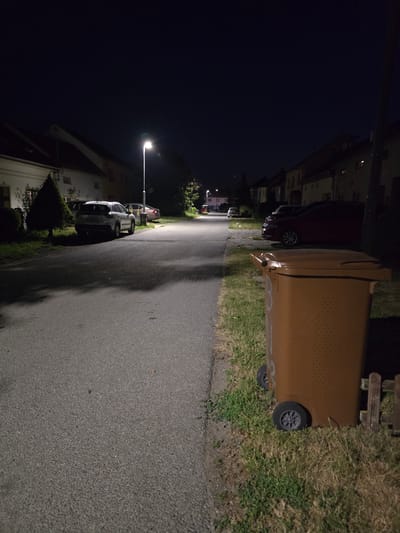 This is a night-time photograph of a residential street in Kostelec na Hané, Czechia. The street is paved and illuminated by a single streetlamp. On the left side of the street, there are houses with pitched roofs and parked cars, including a white SUV and a darker car. A well-maintained evergreen tree is also visible. On the right side of the street, a large brown trash bin with wheels is positioned on a grassy verge next to a wooden fence. Beyond the trash bin, more houses are visible, some with lit windows. The overall scene depicts a quiet, suburban environment after dark.