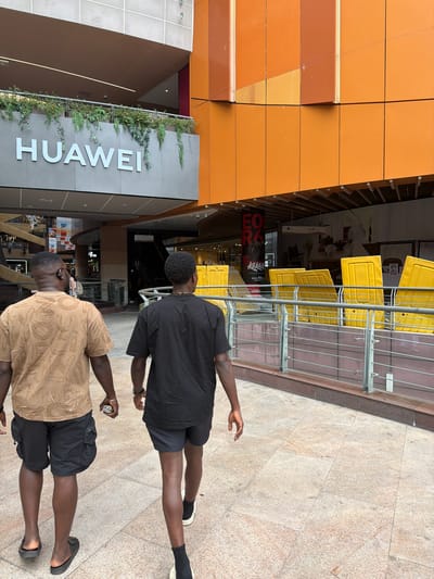 Two Black men are walking away from the camera in an outdoor pedestrian area. The man on the left is wearing a tan t-shirt with a patterned design and black cargo shorts, and black slides. The man on the right is wearing a black t-shirt and black shorts, black socks, and white sneakers. The man on the left is holding a small silver device in his right hand.

Behind them, the environment appears to be a modern commercial district. To the left, a building with a prominent "HUAWEI" sign is visible, with greenery cascading over a railing above it. To the right, a section of a building with large orange panels is present. In the background, there are indications of a shopping mall or commercial complex with escalators, railings, and various storefronts. Yellow protective barriers are lined up along a walkway. The ground is paved with light-colored stone tiles. The scene suggests a public space in a developed urban area.