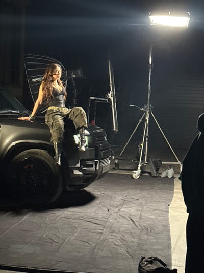 The image captures a nighttime or studio photography session in Ivry-sur-Seine, France. A young adult female, with long wavy brown hair, is prominently featured seated on the front fender and hood of a dark, modern sport utility vehicle, identifiable as a Land Rover Defender. She wears a black strapless corset, olive green cargo pants, and dark boots. Her left arm rests on the vehicle's hood, while her right leg is bent, with her boot positioned near the vehicle's front bumper. The vehicle's left headlight is partially illuminated.

The scene is lit by professional photographic equipment, including a large, octagonal softbox bearing "NANLUX" branding, positioned behind and to the left of the woman. To the right, a tall light stand supports a horizontal rectangular LED panel, providing additional illumination. Other light stands, a silver reflector, and sandbags are visible on the ground. The floor is covered by a wrinkled black fabric extending from beneath the vehicle into the foreground. In the lower right corner, the dark, out-of-focus silhouette of a person, likely a crew member or photographer, is visible, facing the scene. A dark bag rests on the black fabric in the immediate foreground. The background is dark, appearing to be a solid wall or garage door.