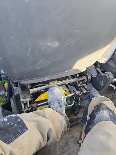 The image displays a first-person, downward-angled view of the interior of a vehicle, in the city of Opalchenets, Bulgaria. The central focus is on a person's lower legs and feet, clad in light brown work trousers with dark grey knee patches extensively covered in white, powdery residue. The person's feet are in dark, dirty work boots. The left boot rests upon a bright yellow object, possibly a rolled-up piece of fabric or tool, situated directly beneath the forward edge of a dark grey vehicle seat. The underside of this seat, visibly soiled with white smudges and streaks, occupies the upper portion of the frame. Exposed black metal seat rails and support structures, heavily coated in dust and grime, are prominent beneath the seat. To the far left, a clear plastic bottle with a red cap is partially visible alongside a yellow reflective garment. The vehicle's floor, seen under the feet, is covered by a dark, dirty mat with visible debris. Sunlight casts a diagonal highlight across the lower right section of the seat and its white trim.