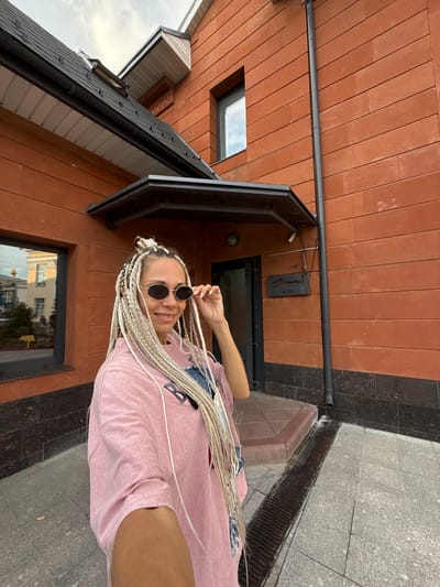 A young woman with blonde braided hair and sunglasses poses for a selfie outside a brick building. She is wearing a pink t-shirt and her hair is long, reaching past her shoulders. The building is made of orange brick and has a dark roof. There is a window to her left and a door with a sign above it to her right. The ground is paved with gray stones, and there is a metal grate in front of the building. The background shows a cloudy sky. The sign on the building is illegible. The building's architecture suggests it might be a shop or restaurant. The overall scene appears to be an outdoor public space in a city.
