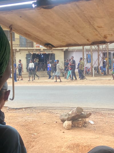 A ground-level, slightly tilted view from within a vehicle or shelter in Okengwe - Obehira, Nigeria, shows a group of people walking along a road. The group comprises primarily young men and boys. They are moving from right to left in the frame along the left side of the paved road. Some are carrying long, thin objects, possibly sticks. The buildings lining the road on the left side have a worn, urban look. Clothes are hung out to dry on balconies or structures attached to the buildings. On the right side of the road, the ground is unpaved with a dirt surface. A pile of logs and rocks sits on the roadside. The view is partially obstructed by the roof of the structure, which has a metallic or corrugated appearance. The lower part of the frame includes a glimpse of an individual wearing green clothing.