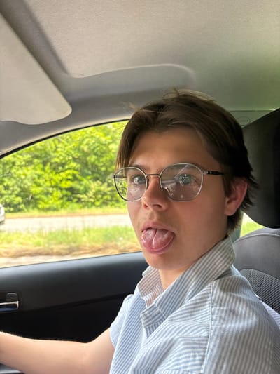 A fair-skinned person in a blue striped shirt and glasses sticks their tongue out. They are in a car, visible by the headrest, car door, and greenery seen through the window. The person faces the camera, with a slight tilt. The glasses are round with thin metal frames. The lighting is bright, possibly midday.