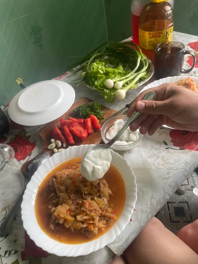 An overhead shot captures a table laden with food, suggesting a meal in progress at an indoor location, likely a home, in the city of Krasnoe, Russia. The table is covered with a white tablecloth patterned with red roses and gold accents. In the foreground, a white bowl holds a stew of chopped cabbage and meat, topped with a dollop of sour cream. A spoon, held by a person's hand, is poised to add more sour cream. Beside the bowl, a small glass dish contains what appears to be cubed white cheese or sugar. To the left, a cutting board displays sliced tomatoes, garlic cloves, and a sprig of dill. Further back, a plate of fresh green lettuce and scallions sits near a dark mug of tea and two bottles, one with red liquid and another amber bottle labeled in Cyrillic. The background features a green tiled wall.