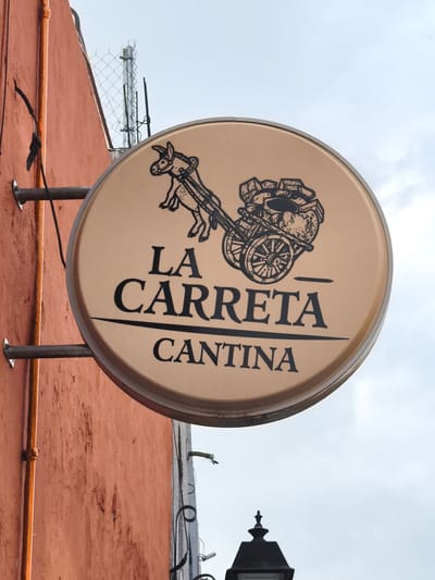 A round, illuminated sign for "La Carreta Cantina" hangs on the side of a red-painted building. The sign is light brown with black lettering and a cartoon donkey pulling a cart. The sign is attached to the building via two horizontal metal bars. The wall is textured and shows signs of wear. A vertical orange pipe is affixed to the wall near the sign. In the background, there is a wire fence and a partly visible street lamp. The sky is overcast. This is likely located in the city of Querétaro, Mexico as indicated by the name of the cantina.