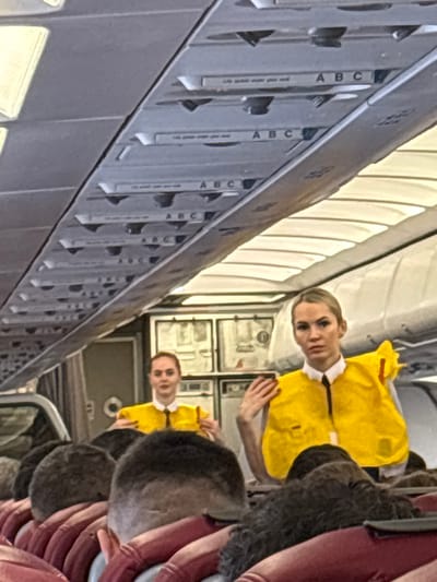 The scene is set within an aircraft cabin in the city of Novogorelovo, Russia. Two female flight attendants are visible in the mid-ground, performing a pre-flight safety demonstration. The flight attendant on the right, with blonde hair, wears a white collared shirt, black tie, and a yellow life vest, demonstrating its use with her right hand on her left shoulder. The flight attendant on the left, with auburn hair, is also in a white collared shirt, black tie, and yellow life vest, seemingly demonstrating another aspect of the vest. Both women face forward towards seated passengers. The foreground shows the tops of multiple passengers' heads and reddish-brown seat headrests. The overhead panels of the aircraft cabin are visible, featuring passenger service units, air vents, and seat row indicators marked "A B C." Text like "life vest under your seat" is faintly discernible on some panels. A galley or service area with closed compartments is visible behind the flight attendants.