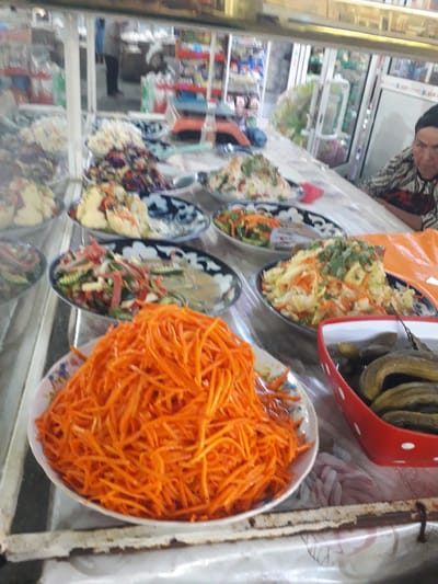 This image captures a vibrant display of salads and pickled vegetables at what appears to be a food counter or market stall in Ghafurov, Tajikistan.

In the foreground, a prominent large bowl is filled with bright orange, finely shredded carrots, characteristic of *Morkovcha* (Korean carrot salad), sitting on a white floral-patterned surface. To its left and further back, several other bowls present a variety of colorful mixed salads, including combinations of shredded cabbage, carrots, green herbs, and possibly other ingredients like tofu or various vegetables. One distinct red plastic container with white polka dots holds a heap of pickled cucumbers or gherkins. Serving utensils are visible in some of the bowls, indicating they are ready for serving.

The scene is set indoors, likely within a shop or market, as suggested by the glass display counter. Reflections are visible on the glass, mirroring parts of the food display and the background. Behind the counter, in the upper left, a person wearing a patterned shirt and a dark head covering is partially visible, suggesting a vendor or worker attending to the stall. Further in the background, blurred shelves stocked with various products and other people or reflections can be discerned, reinforcing the commercial setting. The lighting appears to be ambient indoor light. No discernible text is clearly visible in the image. The arrangement and types of food suggest a focus on fresh or prepared side dishes commonly found in Central Asian cuisine.