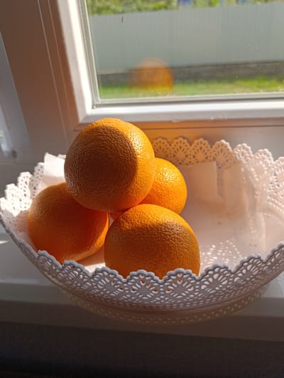 A still life photograph shows four oranges arranged in a white, decorative basket. The basket has a lacy, perforated edge. The oranges are a vibrant orange color and appear plump and textured. Three of the oranges are partially visible, stacked in a way that one is on top of another, and a fourth is to the side. The basket and oranges are placed on a windowsill. Sunlight streams in from the left, casting strong shadows and highlighting the texture of the oranges. Through the window, a blurry background is visible, showing green foliage and what appears to be a white fence, suggesting an outdoor setting. The time of day appears to be morning or late afternoon due to the angle of the sunlight. The scene is peaceful and still, with a focus on the simple beauty of the fruit bathed in natural light. The location context provided is Marshyntsi, Ukraine. There is no visible text in the image.