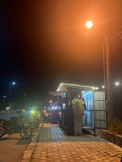 At night in Meunasahaluedrienlhoksukon Barat, Indonesia, a street scene unfolds with a row of parked motorcycles along a paved road. Adjacent to the road is a hexagonal-patterned sidewalk leading to a small open-air food stall. The stall features a corrugated metal structure with a translucent roof illuminated by white LED string lights. Inside the stall, several people are seated, suggesting it is open for business. A light-colored fabric, possibly a tarp or tablecloth, is draped over a display rack at the front of the stall. Further down the street, additional streetlights and a utility pole with wires are visible. The sky is dark, illuminated by a bright, square street lamp casting an orange glow.