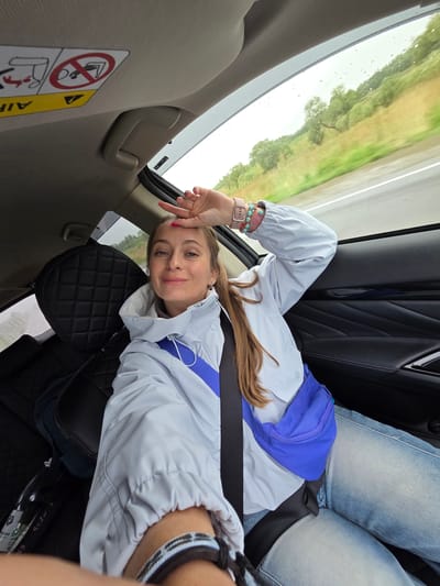 A young woman is captured lying on her back across the rear seats of a car, looking directly at the camera with a gentle smile. She has light brown hair and is wearing a light grey or light blue hooded jacket, blue jeans, and a bright blue fanny pack/sling bag strapped across her. Her left hand is raised to her forehead, revealing pink nail polish, a light pink smartwatch, and several bracelets on her wrist (some beaded, some with blue accents).
The car's interior features dark, possibly quilted, upholstery on the seats and headrest, contrasting with the lighter beige or grey color of the interior ceiling and door panel. A black seatbelt is visible across her torso.
Through the car window, a blurred green landscape of trees and what appears to be a road or field can be seen, indicating the vehicle is in motion. Numerous raindrops are visible on the window, suggesting rainy weather or recent rainfall. The scene is brightly lit, consistent with daytime.
Notable details within the car include a beige grab handle above the window and a partially visible warning label on the car's interior pillar. The label features a red circle with a diagonal line over an icon depicting a child in a rear-facing car seat, and a yellow triangle with an exclamation mark alongside the word "AIRBAG."
The overall impression is a relaxed moment during a car journey in Бабстово, Russia.