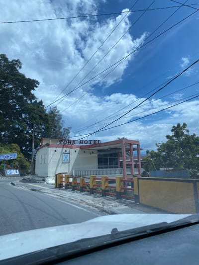 The image shows a street view in Parapat, Indonesia, featuring the "TOBA HOTEL." A white building with red accents is visible, displaying the hotel's name in red lettering on the facade. A smaller logo for Toba Hotel is also present on the building. A white railing with red and yellow supports lines the walkway in front of the hotel. Trees and a blue sky with white clouds are in the background, along with overhead power lines. The street in the foreground appears to be asphalt and shows some signs of wear.