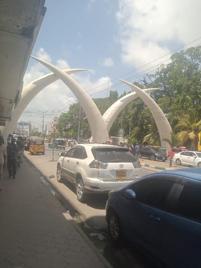 The image depicts a bright daytime street scene in Mombasa, Kenya, centered around the iconic Moi Avenue Tusks monument, an entry archway consisting of two pairs of large, intersecting white structures resembling elephant tusks. This landmark is situated within the city's central business district.
In the foreground, a gray patterned sidewalk extends along the left edge, occupied by several pedestrians walking towards the background. A person in a light-colored top and another in a dark blue uniform-like shirt and dark trousers are visible on the sidewalk. Adjacent to the sidewalk, a white Toyota Harrier SUV (license plate KCH 909V) is parked, facing away from the viewer, with its right wheels positioned in a shallow, water-filled gutter. To its right, a blue hatchback car is partially visible, also parked facing away.
The multi-lane asphalt road stretches into the background. A yellow tuk-tuk is seen driving in the mid-ground, moving away from the viewer, with a small green road sign nearby. Numerous other vehicles, including cars and possibly motorcycles, are present on the road and parked along the sides, indicating active traffic. Buildings, some multi-storied, line both sides of the street in the distance. Lush green trees and palm trees are prominent behind the monument and buildings. The sky is largely clear blue with scattered white clouds, and bright sunlight casts shadows across the scene. Utility poles and overhead power lines crisscross the upper portion of the frame. The overall environment suggests a busy urban commercial area.