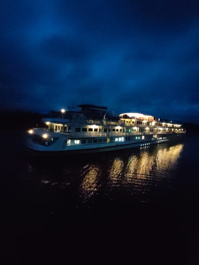 A passenger ship is docked in the city of Devyatiny, Russia. The ship is white with multiple decks and illuminated windows, reflecting its lights onto the dark water. The sky is a deep blue, suggesting it is either dusk or dawn. The ship appears to be the focal point of the image, with the surrounding environment largely obscured by darkness.