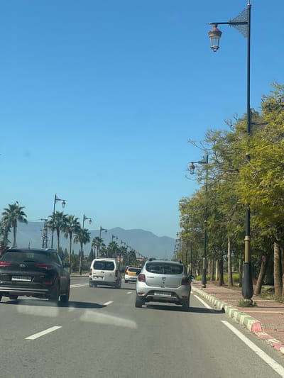 A daytime street scene in Beni Mellal, Morocco, is depicted from a forward-moving vehicle. The asphalt road features white lane markings, including a prominent white arrow indicating straight travel. Vehicular traffic ahead includes a black Hyundai Tucson SUV (partially visible rear, foreground left), a silver Dacia Sandero hatchback (foreground right), a white panel van (mid-ground left), and a yellow car (mid-ground right).
To the right of the road, a paved sidewalk is bordered by a curb painted in alternating white, green, and pink sections. Along this sidewalk, a row of mature trees with green foliage stands, interspersed with ornate, dark-colored streetlights featuring classic lantern-style fixtures.
To the left, a planted median separates traffic lanes, containing numerous tall palm trees and similar decorative streetlights. Utility poles with overhead lines are visible among the palm trees. In the distant background, under a clear, bright blue sky, large mountains are visible.