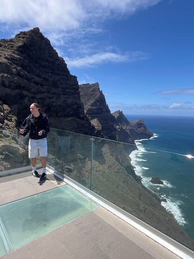 The image shows a man standing on a glass platform with a dramatic mountain and ocean backdrop, likely at the Mirador del Balcón (also known as Mirador de Anden Verde) in La Aldea de San Nicolás, Spain. The man, dressed in a black jacket and white shorts, is the focal point. Behind him are steep, dark cliffs that plunge into the ocean, where waves are visible crashing against the rocks. The sky is blue with some wispy clouds. The glass platform is elevated above the landscape, creating a sense of exposure and height.