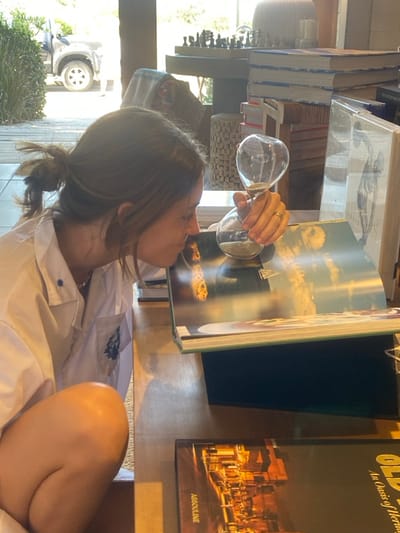 A young woman holds an hourglass to her mouth, its contents appearing to flow into an open book. The book features a nautical scene of sailboats on the ocean under a partly cloudy sky. Beside the book is another titled "Old Oasis of Heritage" by Assouline, depicting a city at night. The setting seems to be a storefront, with books stacked in the background and a glimpse of a car through the window. The woman is wearing a white collared shirt.