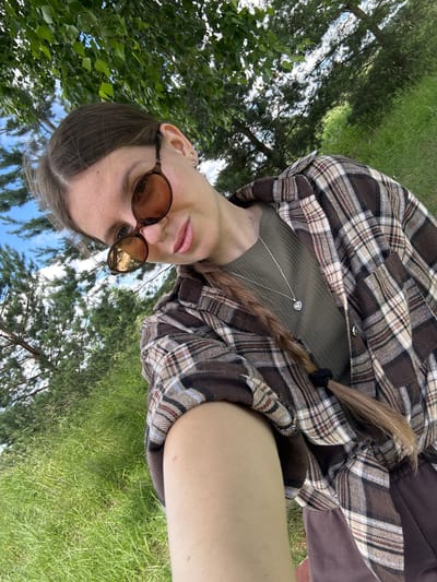 A woman is taking a selfie outdoors. She is wearing sunglasses, a plaid shirt, and a green top. Her hair is in a braid. The background shows trees and green grass, suggesting a natural, possibly park-like setting. The lighting indicates it is daytime, likely with sun. The specific location in Borshchevka Malaya, Russia, is not discernible from the visual information provided in the image.