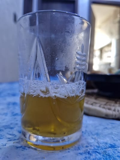 A clear, faceted glass containing a yellowish-brown liquid with white foam and dark sediment at the bottom is positioned centrally on a mottled blue and white surface in the city of Saâda, Morocco. The glass exhibits condensation on its exterior, and molded patterns are visible on its sides, including vertical lines and a stylized leaf or feather motif. Small bubbles adhere to the inner surface of the glass above the liquid line. The blue and white surface, possibly a table or tablecloth, has scattered small crumbs or debris. The background is out of focus, revealing a light-colored wall with a darker vertical element on the left, and a brighter, indistinct area suggesting an opening or light source on the right. A dark, blurry object is also visible in the far-right background. No individuals are present in the image.