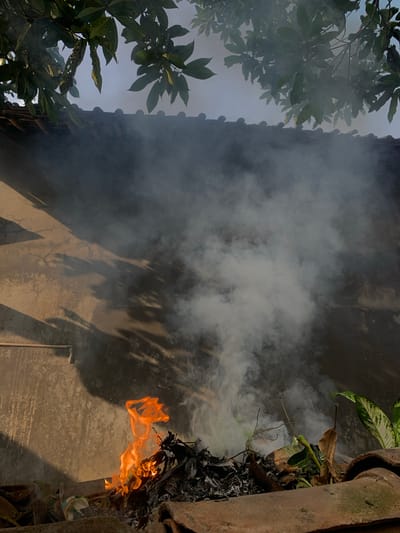 A fire burns amongst debris on what appears to be a rooftop in Karangkemiri, Indonesia. Flames, primarily orange and yellow, erupt from a pile of dried leaves, branches, and other organic matter. White smoke billows upwards, obscuring a textured, tan-colored wall with shadows cast upon it, possibly from nearby foliage. The wall is punctuated by a simple horizontal pipe near the bottom left. Above the wall, a portion of a dark-tiled roof is visible, and overhanging green leaves from a tree create a canopy against a hazy sky. The scene is captured from a low angle, focusing on the fire and smoke in the foreground.