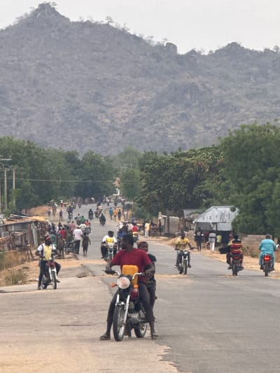 A street scene in Dawaki, Nigeria shows a large group of people riding motorcycles on a paved road. The road is lined with trees and small buildings. A mountain range is in the background. Many people are alongside the road.