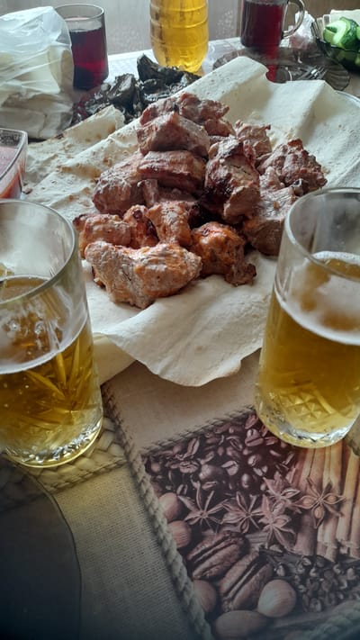 The image depicts a close-up, overhead view of a table laden with food and drinks, likely in a domestic setting in Bagayevskaya, Russia. The focal point is a platter of cooked meat, appearing to be shashlik or kebabs, served on a bed of flatbread. Two glasses filled with a pale yellow, effervescent beverage, presumably beer, are prominent in the foreground. To the left, a glass of dark red liquid, possibly wine or juice, is visible alongside a partially opened plastic bag. In the background, there are other containers with food items, including what looks like a bowl of sliced cucumbers. The table surface is covered with a patterned cloth featuring images of coffee beans and spices. The lighting suggests an indoor environment, possibly during daylight hours. There are no individuals visible in the image.