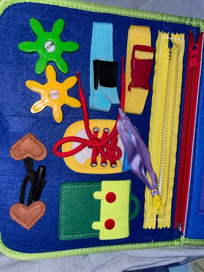 This media file is a close-up shot of a blue felt busy board, likely a toy for a child. The busy board is filled with various interactive elements designed to stimulate fine motor skills and sensory exploration.
Dominating the frame are several objects attached to the felt. At the top, there's a red zipper with a silver pull tab. Below it, a yellow zipper with a yellow pull tab is partially open, revealing its teeth. To the left of the yellow zipper, a yellow strap is fastened with a red plastic buckle. Below this, a light blue strap with a black plastic buckle is also visible.
A prominent feature is a yellow felt cutout resembling a shoe, complete with eyelets and a red shoelace threaded through it and tied in a bow. A purple ribbon is also tied to the shoelace.
To the right of the shoe cutout is a light green felt backpack shape with two red circular embellishments that could represent buttons or pockets.
In the bottom left quadrant, two colorful star-shaped plastic toys are attached to the felt. The one on the left is green with a white center, and the one on the right is yellow with a white and silver center.
In the bottom right quadrant, two small brown felt hearts are attached to a black cord with a black toggle clasp.
The background appears to be a light-colored fabric, possibly bedding, suggesting the busy board is laid out on a bed or a soft surface. There is no visible text in the image. The lighting is even, indicating an indoor setting, likely during the daytime. The overall impression is of a playful and educational toy designed for young children, with no specific time of day, weather, or emotional cues evident. The context provided indicates the location as Gamovo, Russia.