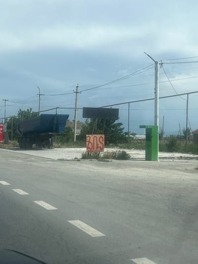 This image presents a roadside scene in Ayntap, Armenia, captured on a bright, clear day. The foreground features an asphalt road with distinct white dashed lane markings. On the left side of the road, a dark blue dump truck is visible, positioned facing away from the viewer.

Numerous utility poles with interconnected power lines run parallel to the road, dominating the midground. One tall, light-colored utility pole stands prominently on the right side of the road. Adjacent to this pole is a distinct bright green utility box or cabinet; faint text, possibly "YASTEL," can be observed on its surface.

Near the dump truck, a small, reddish-brown sign displays the red Armenian letters "3ԱՏ," which transliterates to "ZAT." The background behind the road and utilities consists of sparse green vegetation, bushes, and some distant structures or fences, indicative of a semi-developed or industrial area. The sky above is light blue with scattered white clouds, suggesting sunny weather during daytime.