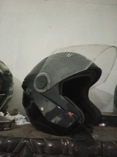 The scene depicts two motorcycle helmets positioned on a dark, textured surface, likely indoors against a light-colored, textured wall in the city of B. Nam Ngao, Laos. The primary subject is a dark-colored, possibly black, open-face motorcycle helmet located in the center-right. Its clear visor is partially raised and shows some accumulation of dust or debris. A dark, circular pivot mechanism for the visor is visible on the left side of the helmet. A dark strap hangs from the bottom of this helmet. To the left, a second motorcycle helmet, partially obscured, is visible; it appears to be multi-toned, possibly dark green or camouflage pattern. Between the two helmets, on the surface, are several pieces of crumpled white tissue or paper. A dark object, possibly a box, with the partial word "BOLD" printed on it, is situated to the left of the main helmet. No people, active actions, or interactions are visible.